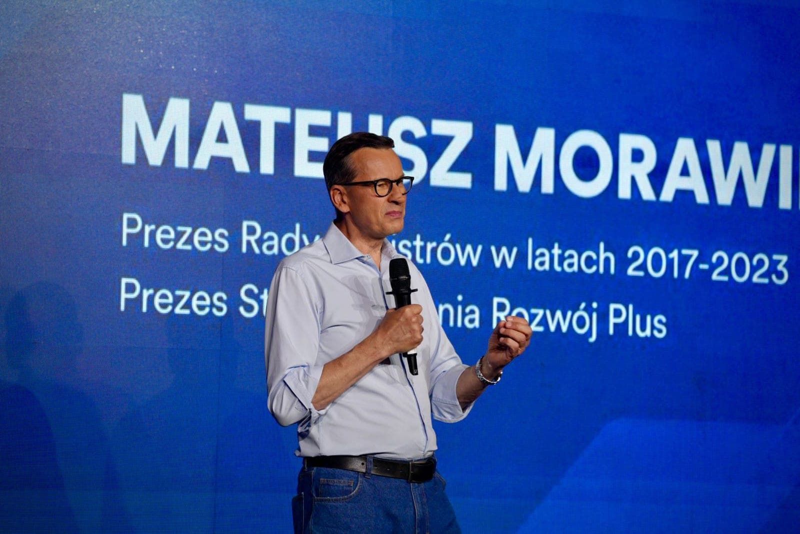Mateusz Morawiecki przemawia z mikrofonem na scenie, za nim niebieski slajd z jego nazwiskiem.