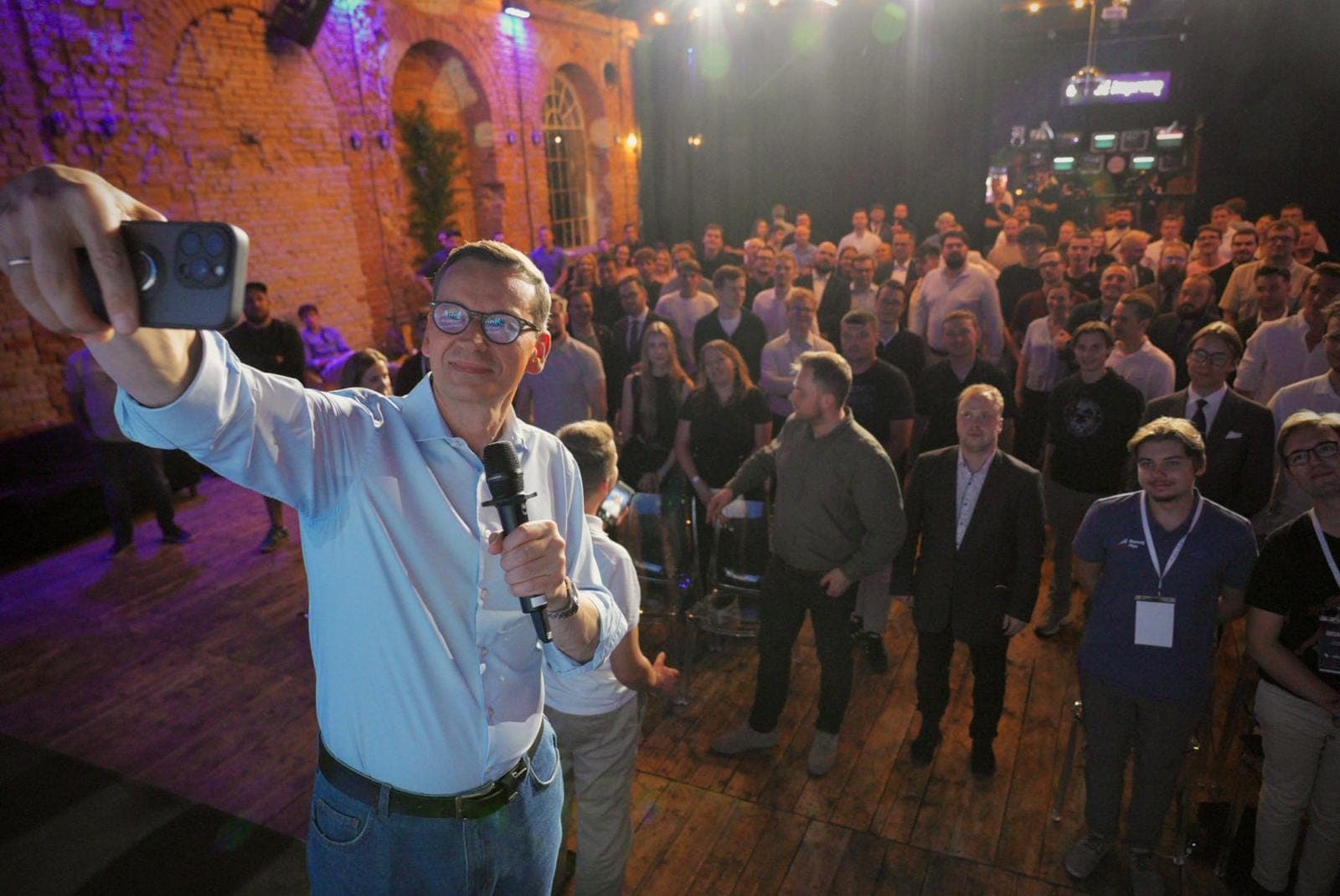 Mateusz Morawiecki robi selfie ze sceny, w kadrze pełna sala uczestników w ceglanej hali.